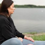 Woman meditating outdoors by a tranquil lakeside, fostering health and wellness.